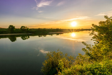 Naklejka na ściany i meble Scenic view at beautiful summer river sunset with reflection on water with green bushes, grass, golden sun rays, calm water ,deep blue cloudy sky and glow on a background, spring evening landscape