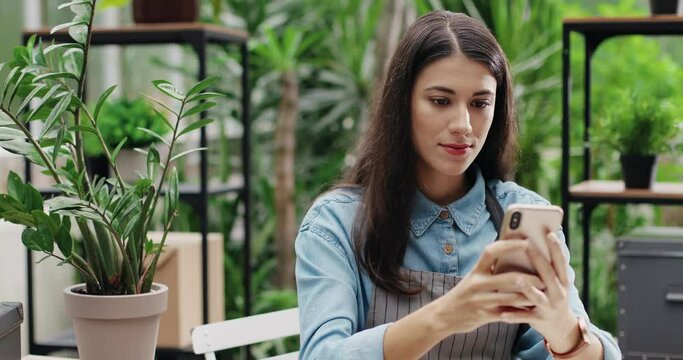 Close up portrait of cheerful Caucasian woman in good mood texting on smartphone in own floral center. Pretty happy female florist at work in flower store typing on mobile phone. Business concept