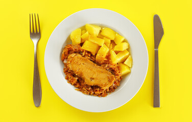 Baked potatoes, pork meat and cabbage in white plate on yellow board with cutlery, view form above
