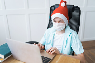 Fototapeta na wymiar A boy in a disposable face mask and Santa Claus hat on Christmas online classes