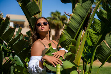 Fashion cute young teenage girl or woman posing with banana tree leaves dressed in summer hipster vintage white stylish clothes. Trendy girl posing. Funny and positive woman in sunglasses