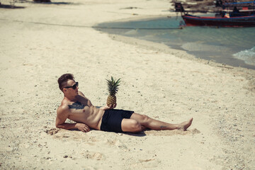 happy shirtless young hipster man  in shorts and sunglasses  with tattoo lying and holding pineapple on sea beach. Summer vacation. Tropical seascape with boats on the background.