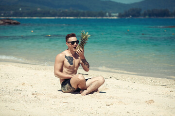 Stylish muscular hipster young man with tattoo wearing sunglasses trying to eat pineapple and sitting on the beach. Summer time concept. Phuket. Thailand