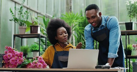 Portrait of happy African American male and female florists flower store owners working and doing inventory. Woman typing on laptop at desk in floral shop and talking to man boss. Business concept