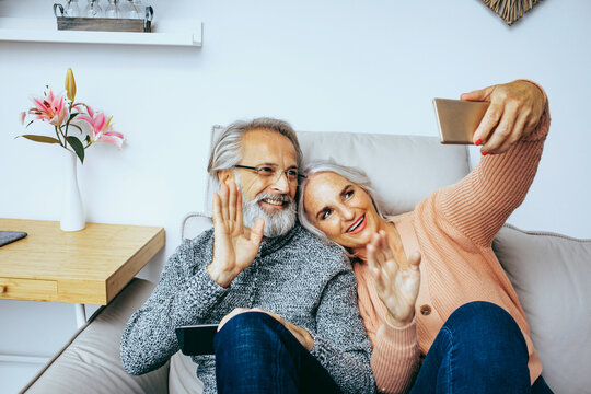 Happy Senior Couple At Home Staying Indoors Connected And Waving To Family Online On Mobile Phone