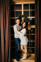 Wonderful couple stand near decorated window, charming husband gently hug beautiful pregnant wife, enjoy tender moments, winter holidays at home, New Year celebration concept