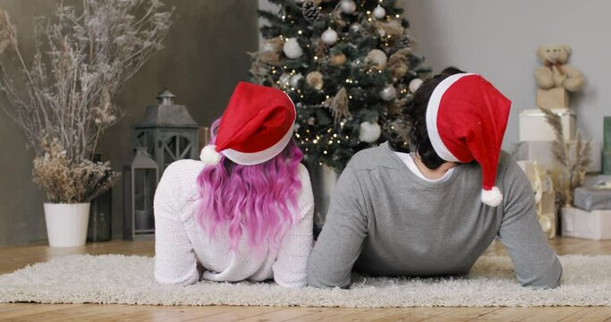 New Year, Christmas, Xmas, holiday and celebration concept. Happy young couple in Santa's red hats lying together near Christmas tree. Family lovely moments. They are kissing and hugging each other.