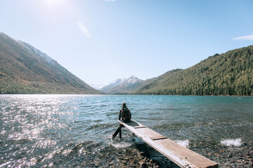 Young hiker wearing blue jacket sitting on the wooden bridge by the mountain lake and looking at the distance