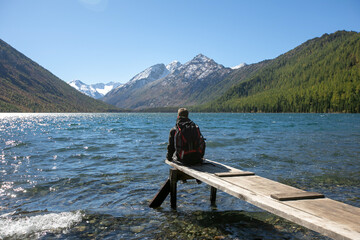 Young hiker wearing blue jacket sitting on the wooden bridge by the mountain lake and looking at the distance
