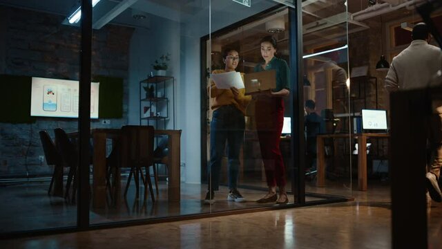 Two Diverse Multiethnic Female Have a Discussion in Meeting Room Behind Glass Walls in an Agency. Creative Director and Project Manager Compare Business Results on Laptop and App Designs in an Office.