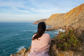 Woman on a pink shirt looking at the landscape of Cabo Espichel Cape sea cliffs and atlantic ocean, in Portugal