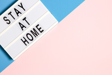 White lightbox with text STAY AT HOME on pale pink and light blue background. Healthcare, medical, quarantine concept. Top view