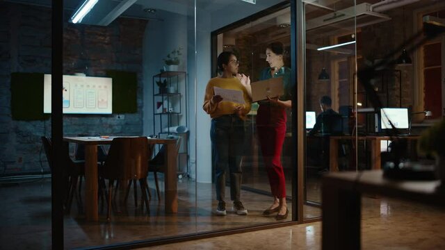 Two Diverse Multiethnic Women Have a Conversation in a Meeting Room Behind Glass Walls in an Agency. Creative Director and Project Manager Talk About Business Results and App Designs in an Office.