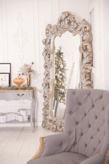 A large carved gray mirror with a reflection of a Christmas tree, a soft gray armchair, a vintage chest of drawers.