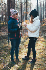 Mother and daughter with backpack having break during autumn trip pouring a hot drink from thermos flask on autumn cold day. Active women wandering in a forest actively spending time