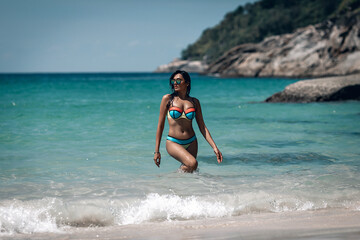Beautiful brunette girl in colorful bikini and sunglass standing in water at exotic beach. Phuket. Thailand. Beautiful blurred seascape