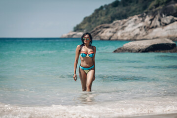 Beautiful brunette girl in colorful bikini and sunglass standing in water at exotic beach. Phuket. Thailand. Beautiful blurred seascape