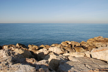 Blue sea stones and clear sky.
Mediterranean Sea, sunny day. In the foreground are textured stones illuminated by the sun. Calm sea with blue water. Surfer at sea. Bright clear sky.