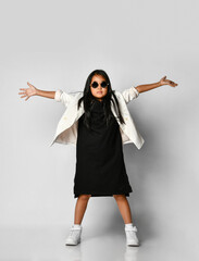 Little Asian girl demonstrates stylish autumn baby clothes, laughing for the camera with outstretched arms. The model is wearing a black dress, white jacket and trendy sunglasses.