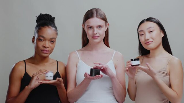 Three healthy looking beautiful females showing moisturizing cream for diverse skin tapes smiling at camera isolated in background. Women beauty. Cosmetics.