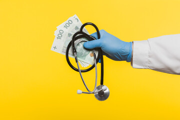 Profile side view close-up of a human hand in blue surgical gloves, holding a stethoscope in hand and showing a bundle of Polish 100 zloty banknotes, isolated on a yellow background.