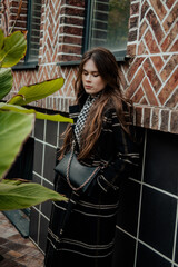 Young attractive woman in trench coat thoughtfully looking in camera while walking around cozy city street