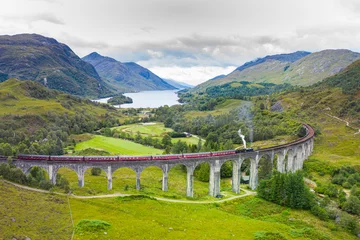 Fotobehang Glenfinnanviaduct Classic Steam Train travelling across an old bridge with dramatic landscape in the background in Scotland 