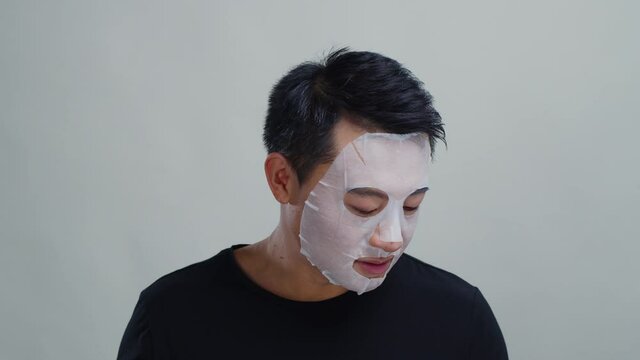 Handsome metrosexual chinese man wearing cleansing face mask skincare product answering phone call talking with girlfriend on grey background isolated.