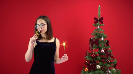 A young woman stands with a glass of champagne and a sparkler near the Christmas tree on a red background. A girl with glasses and an evening dress.