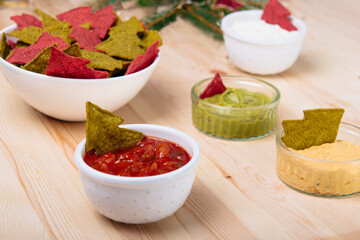 Christmas festive snacks for the party. Green and Red Tortilla chips in the shape of a Christmas tree in bowls with salsa, guacamole, cheese, and mayo sour cream dipping sauces on the wooden table.