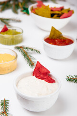 Christmas festive snacks. Green and Red Tortilla crisps in shape of Christmas tree in bowls with sour cream, salsa, guacamole, cheese dipping sauces on the white table decorated with spruce. Vertical.