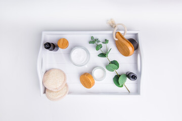 Bamboo face brush, loofah pads, cosmetic natural oil serum, handmade creams and eucalyptus on wooden tray. DIY home eco-friendly spa, massage, peeling and hydration products. Top view. Copy space.