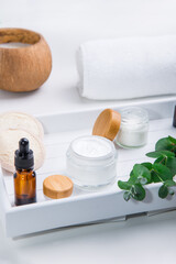 Herbal cosmetic creams in opened containers, fresh eucalyptus, loofah pads and cosmetic natural oil serum on a white wooden tray. Set for face self-care and DIY home eco-friendly hydration. Vertical.