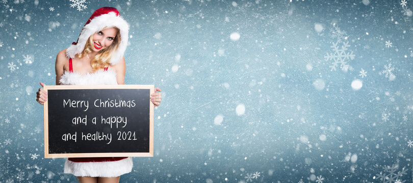 smiling miss santa holding blackboard with message MERRY CHRISTMAS AND A HAPPY AND HEALTHY 2021 in front of winter background