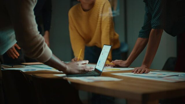 Close Up of Diverse Multiethnic Team Having Conversation in Meeting Room in a Creative Office. Colleagues Lean On a Conference Table, Look at Laptop Computer and Make Notes with Pencils on Notebooks.