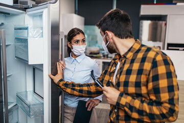 Fototapeta premium Young man talking with saleswoman about refrigerator he wants to buy. They are both with face protective masks. Pandemia, Covid-19 concept.