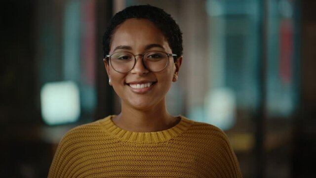 Close Up Portrait of a Young Latina with Short Dark Hair and Glasses Posing for Camera in Creative Office. Beautiful Diverse Multiethnic Hispanic Female Wearing Yellow Jumper is Happy and Smiling.