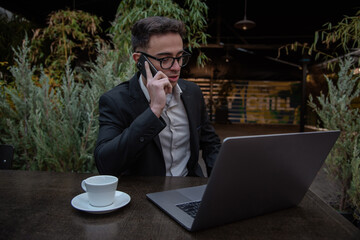 Young successful businessman working on a laptop while sitting in coffee bar during work break lunch. Speaking on his phone while working and drinking coffee.	