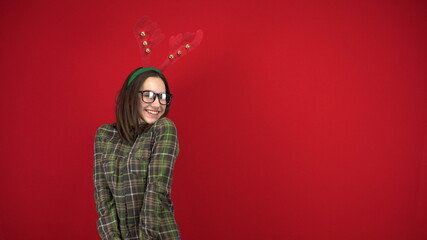 A young woman stands with a headband in the form of Christmas horns and is shy. Studio photography on a red background.