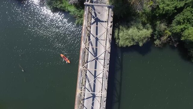 Top Down View Of Metal Bridge Over The River Veleka, While A Car Is Going Through. Two People Are Paddling On A Kayak Through The River.