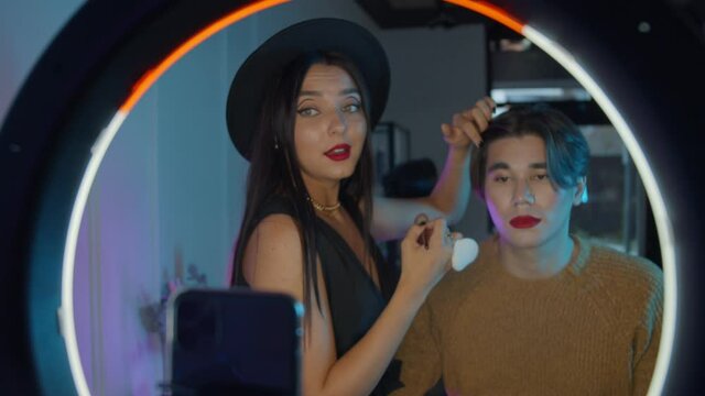Makeup studio - young handsome man with bold red lipstick and his make up artist standing in front of a ring light - adding finishing touches