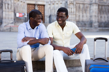 Portrait of two positive african american tourists with luggage on bench of european city