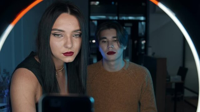 Makeup studio - young handsome man with bold red lipstick and his woman make up artist standing in front of a ring light - adding finishing touches