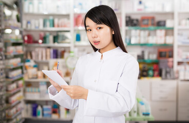 Chinese female looking medicine with notebook near shelves in pharmacy. High quality photo