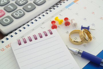 Planning wedding concept. Golden wedding rings, push pins, notepad, pen and calculator on calendar.