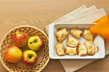 Hot baked in the oven slices of sweet apples in a ceramic form and fresh apples in a wicker basket on a wooden table. Delicious healthy homemade food. Organic products. Flat lay, copy space, close-up