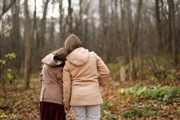 Fototapeta premium Caucasian children sisters walk together in the park in the forest along the path, girlfriends children hold hands and hug on a walk, view from the back, girly secrets, autumn walks in warm coats