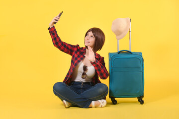 Young girl with short hair in a red checked shirt and blue jeans sits next to a blue travel suitcase and talks to someone online on the phone on a yellow background: place for text, a tourist concept