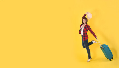 A young happy girl with short hair, a straw hat, a red checked shirt and blue jeans walks with a blue travel suitcase on a yellow background: a place for text, a tourist concept, travel banner