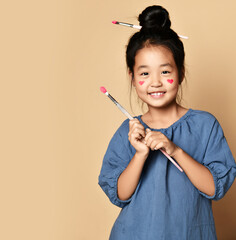 Cute smiling asian kid girl artist with painted red hearts on cheeks and with brush in hair and in hands over yellow wall background with copy space. Trendy children fashion, asian outfit concept